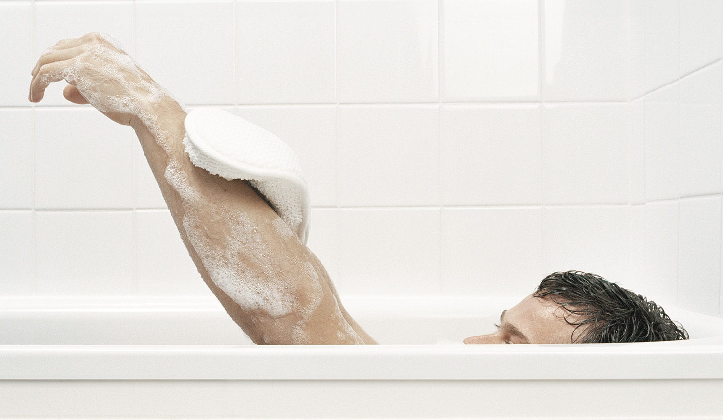 Hombre lavando su brazo con esponja y espuma durante el baño, mostrando una rutina de higiene y cuidado de la piel.